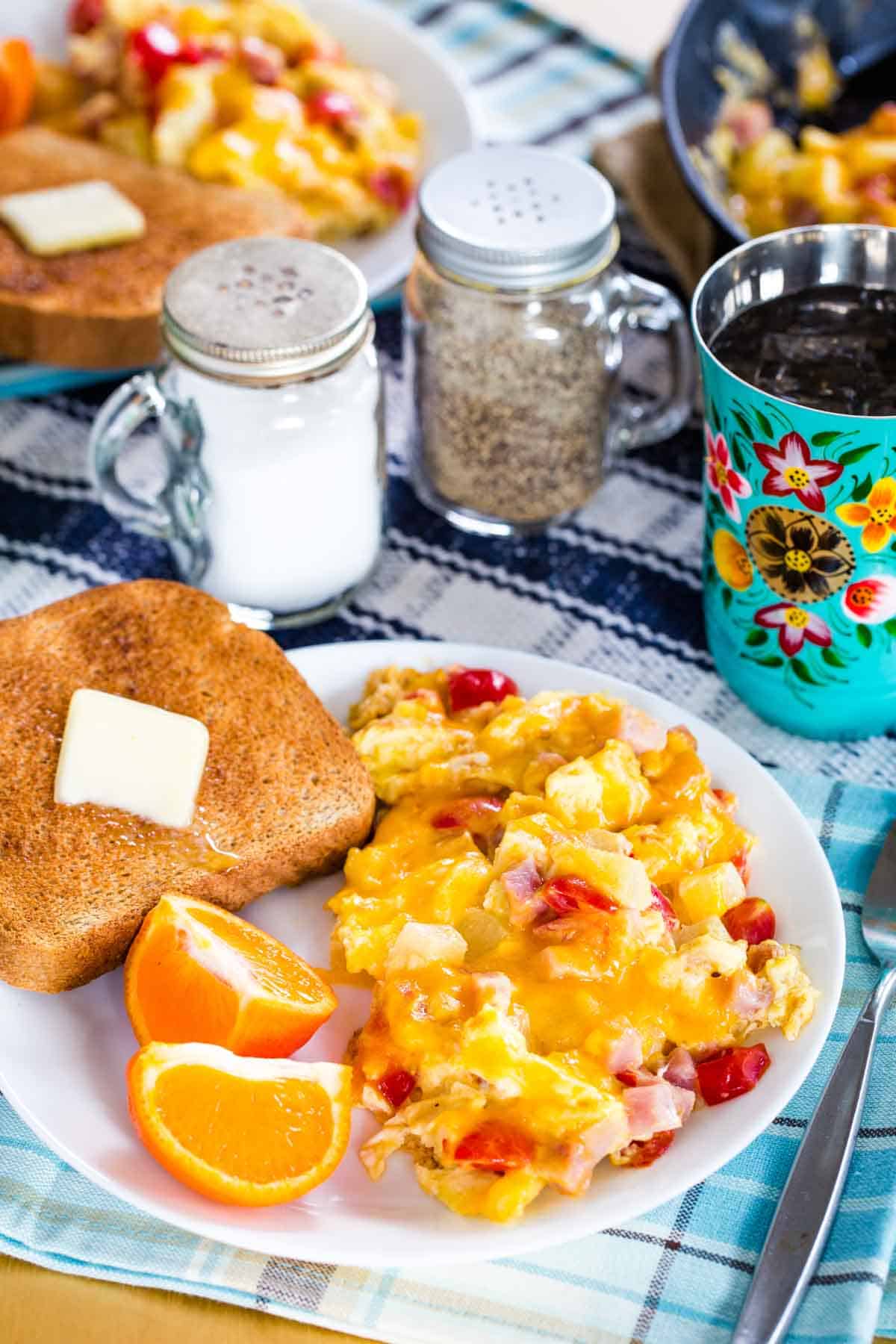 A plate of scrambled eggs, oranges, and toast on a tale set for breakfast with a form, cup, and salt and pepper shakers.