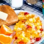 A scoop of cheesy scrambled eggs with ham and tomatoes on a plate alongside orange wedges and a piece of toast with a pat of butter.