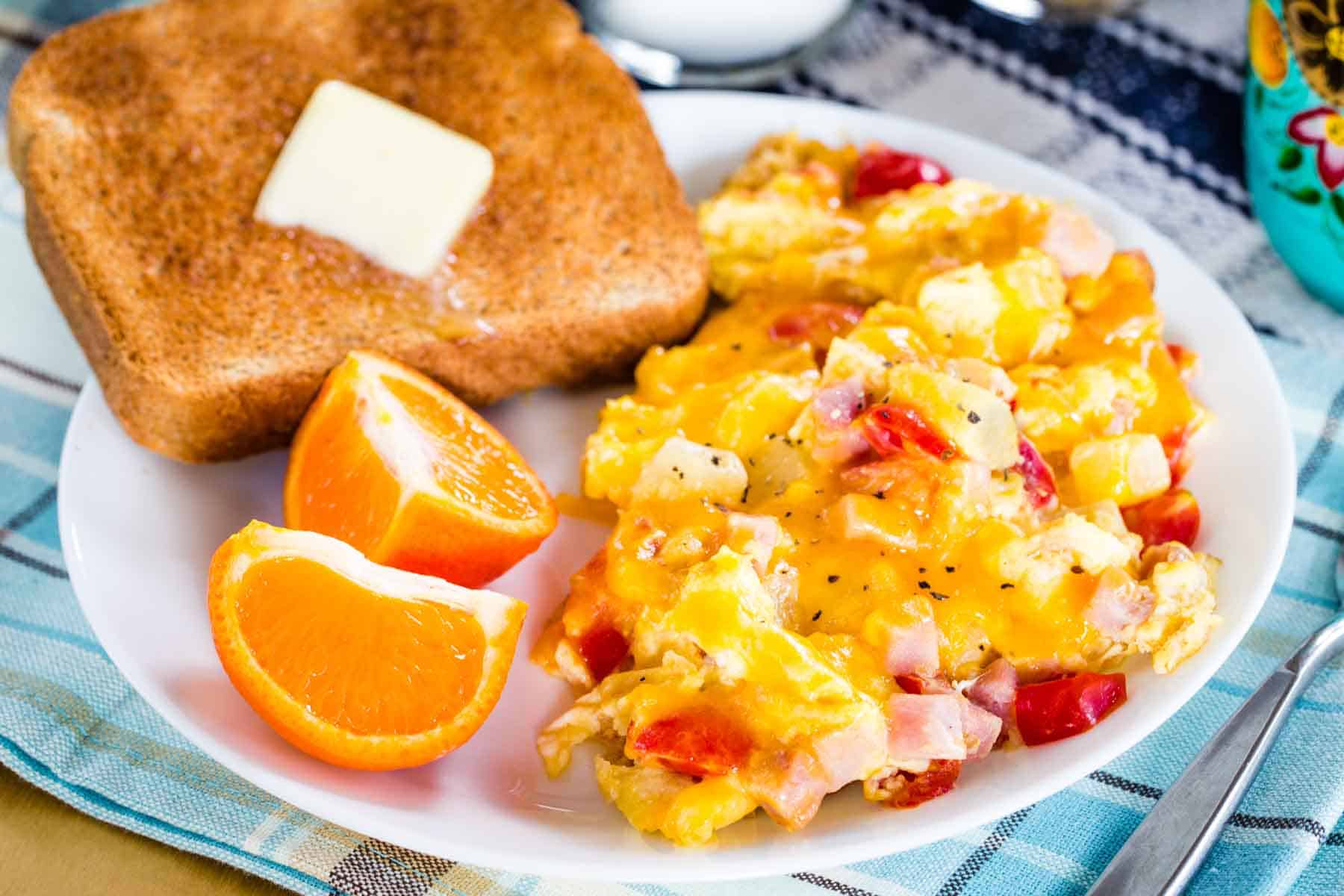 A plate of scrambled eggs with ham, cheese, pineapple, and tomatoes served with toasted and wedges of an orange.