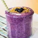 Blueberry Cheesecake Smoothie garnished with graham cracker crumbs and blueberries on top with a green and white straw in a glass