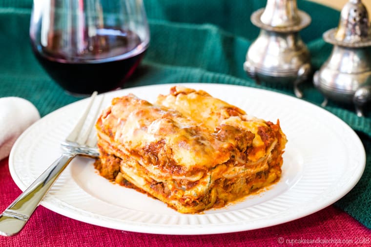 The World's Best Gluten Free Lasagna on a plate on top of a red placemat and a green cloth napkin with a glass of red wine and fancy salt and pepper shakers.