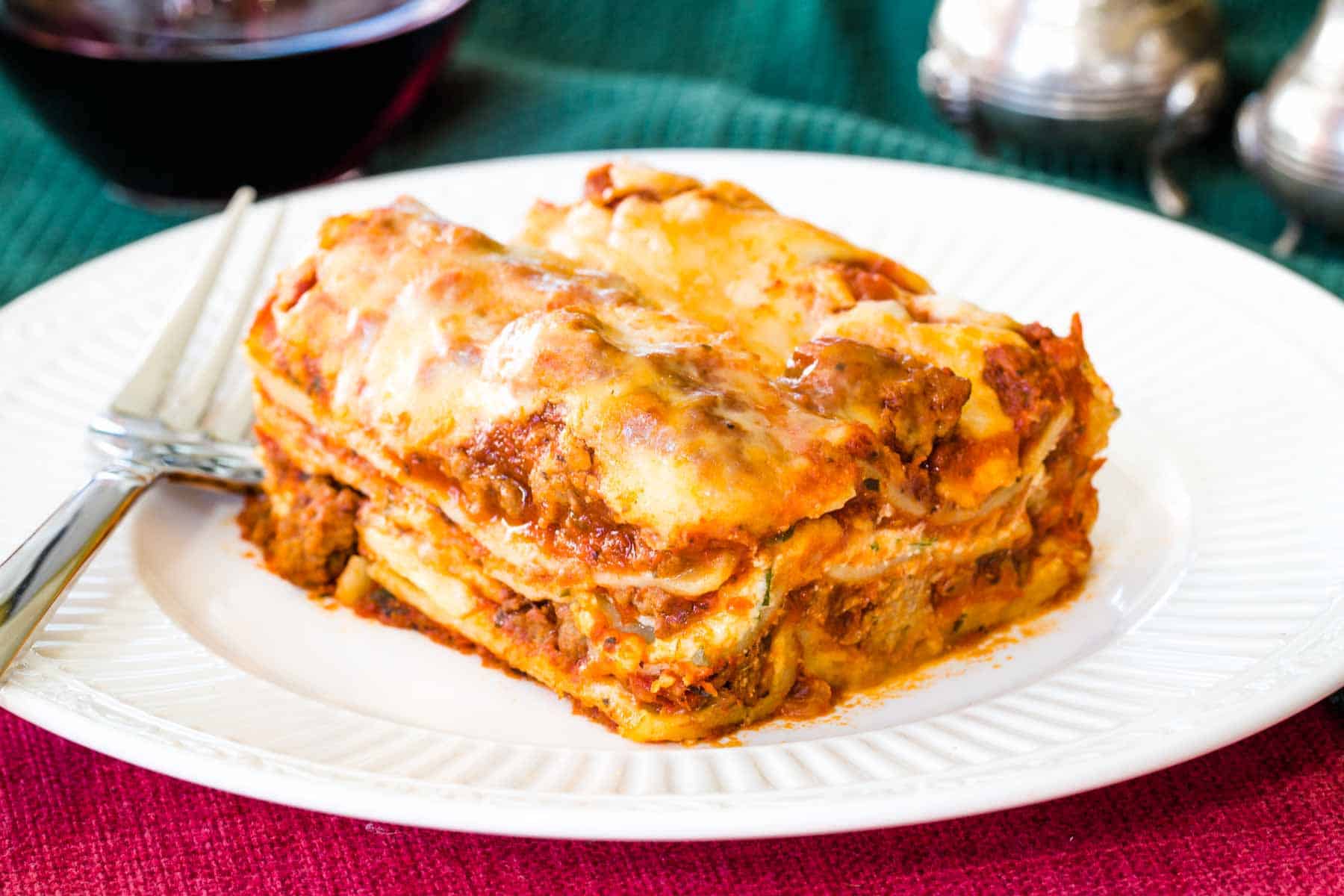 The World's Best Gluten Free Lasagna on a plate on top of a red placemat and a green cloth napkin with a glass of red wine and fancy salt and pepper shakers.