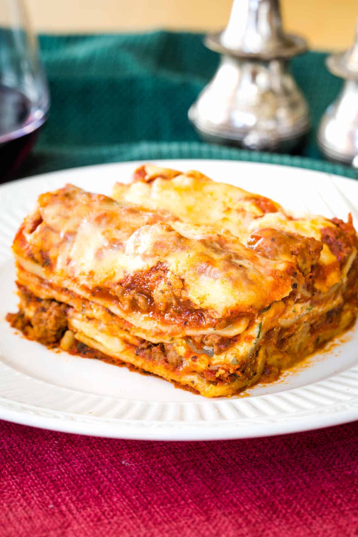 A slice of Gluten Free Lasagna on a white plate set on top of a red placemat with a green cloth napkin behind it.