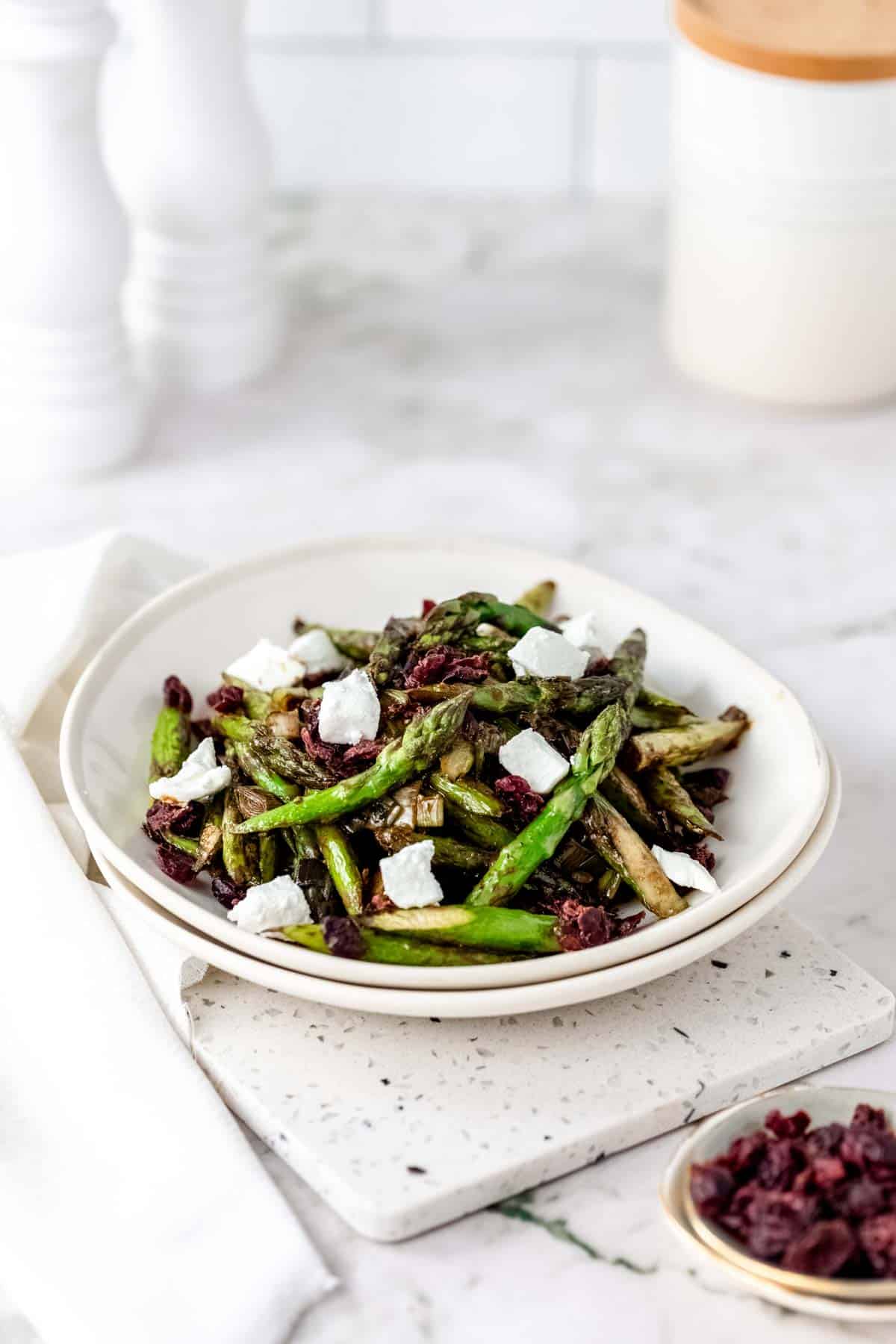 Roasted asparagus salad in a white bowl topped with chunks of goat cheese and dried cherries.