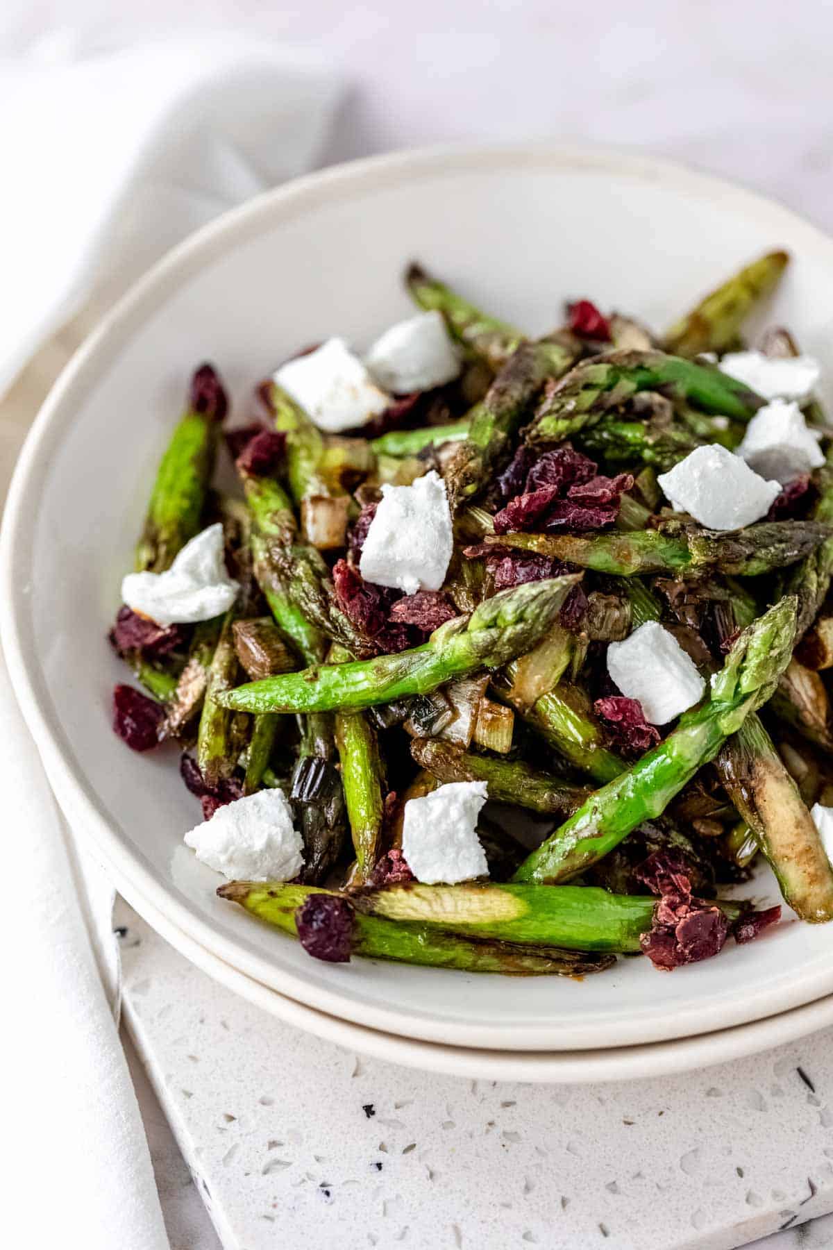 Roasted asparagus salad in a white bowl topped with chunks of goat cheese and dried cherries.