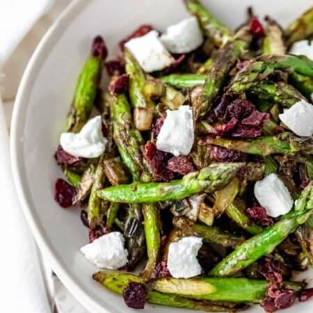 Overhead view of roasted asparagus salad in a white bowl topped with chunks of goat cheese and dried cherries.