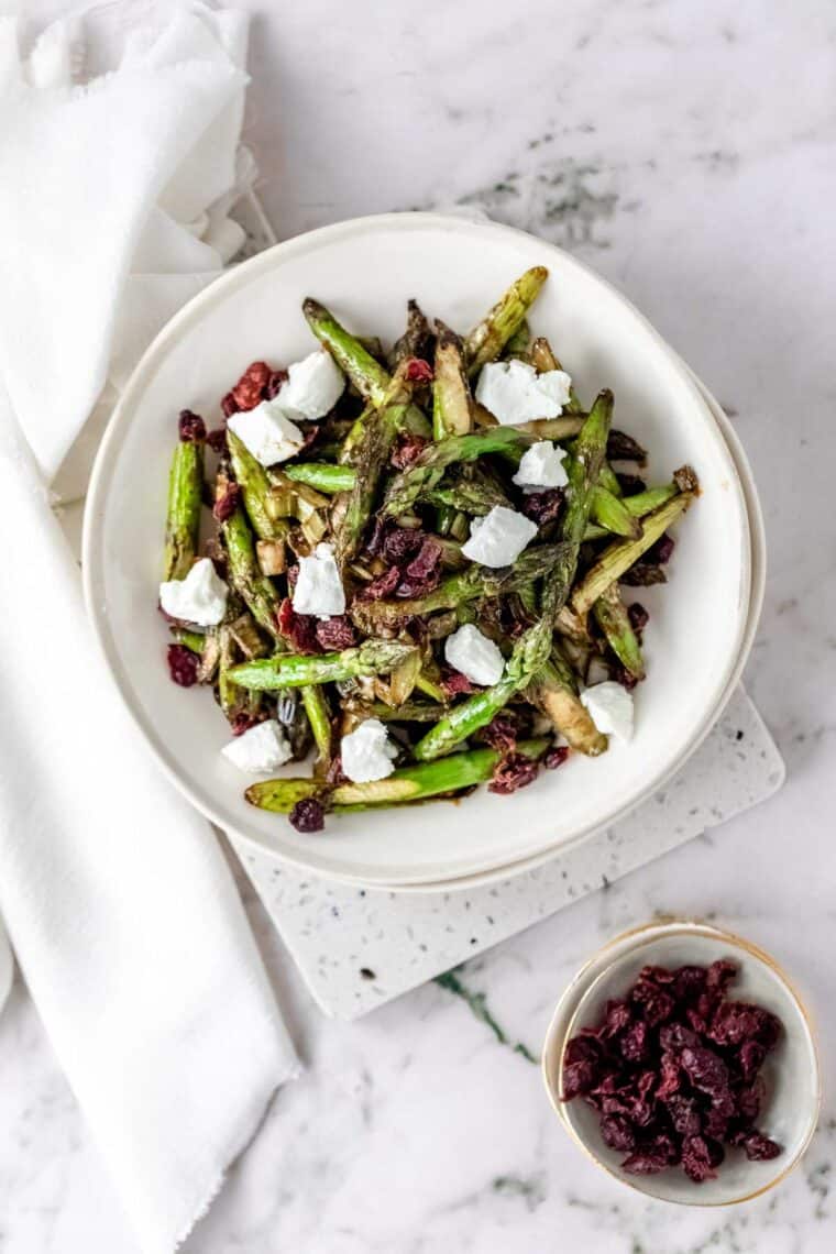 Overhead view of roasted asparagus salad in a white bowl topped with chunks of goat cheese and dried cherries, next to a small bowl of dried cherries.