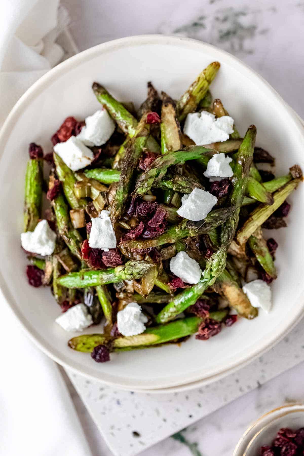 Overhead view of roasted asparagus salad in a white bowl topped with chunks of goat cheese and dried cherries.