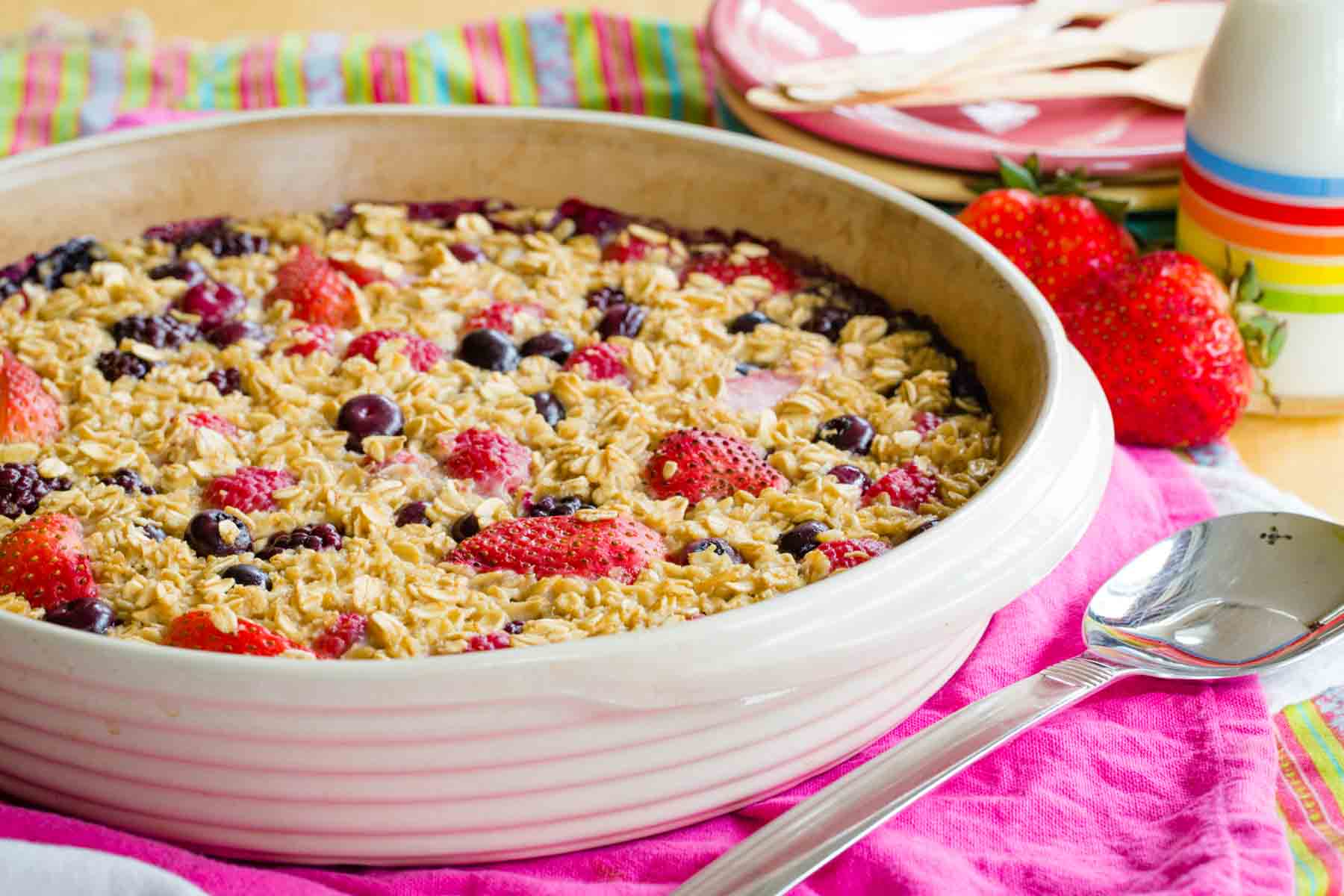 Round baking dish with Mixed Berry Baked Oatmeal on colorful cloth napkins with a stack of plates and a small bottle of milk in the background.