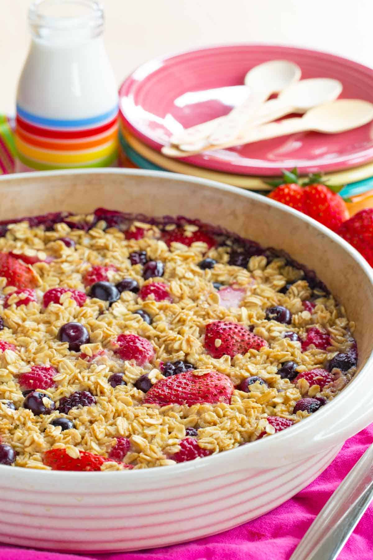 Mixed Berry Baked Oatmeal with a baking dish with a stack of different colored plates in the background with spoons on top of the plates.