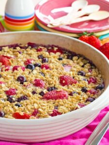 Mixed Berry Baked Oatmeal with a baking dish with a stack of different colored plates in the background with spoons on top of the plates.