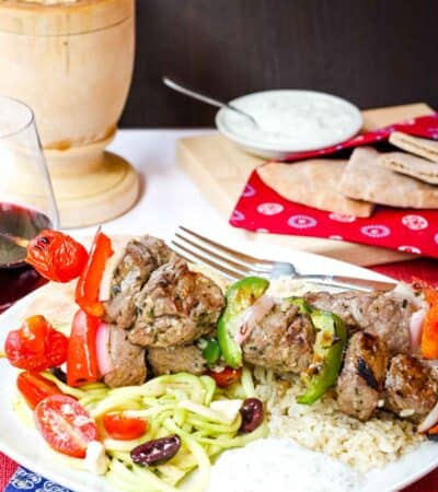 Grilled Greek Beef Kabobs on a dinner plate with rice, hummus, Zoodles Greek salad, and tzatziki on top of a red placemat and a blue cloth napkin with white flowers.