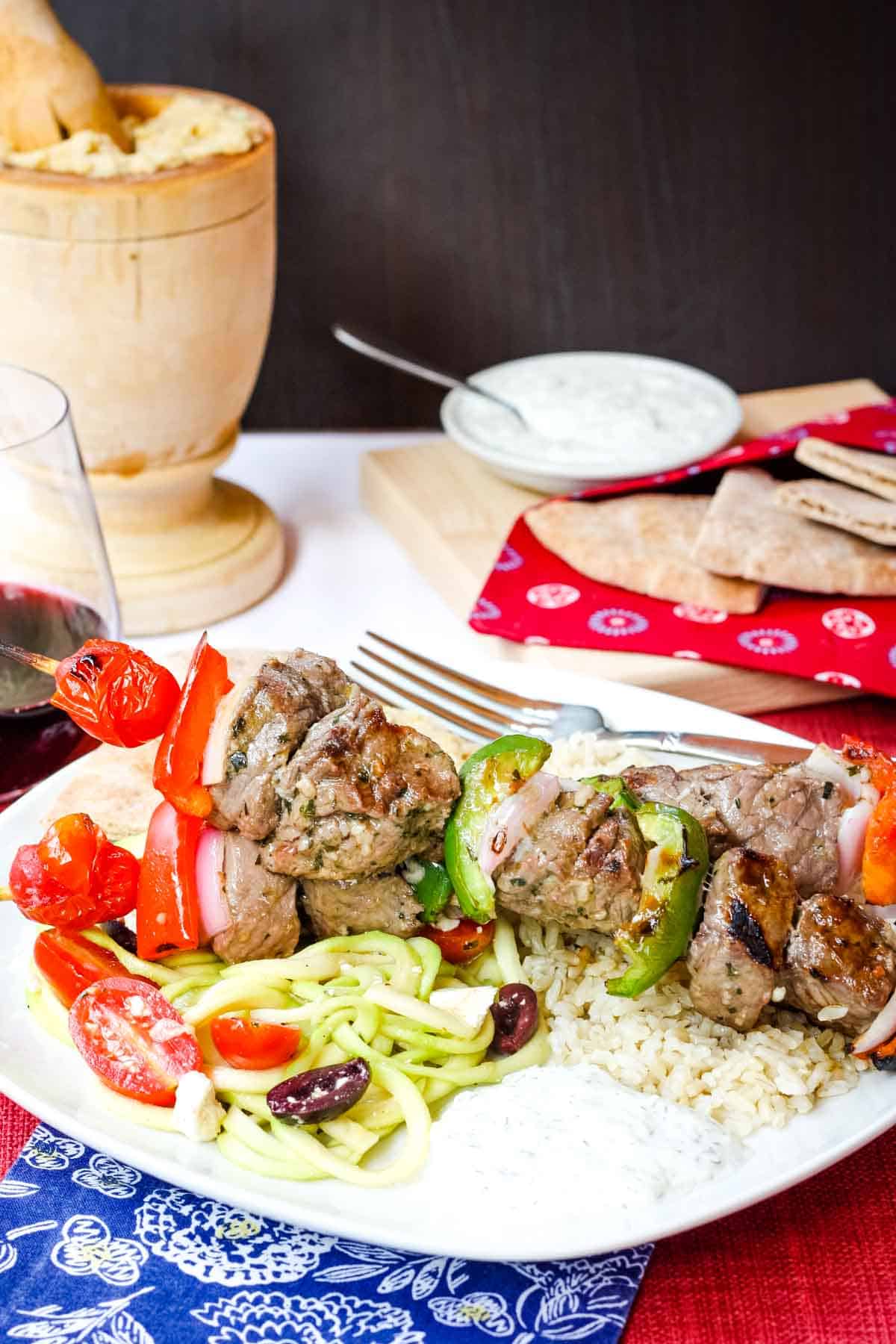 Grilled Greek Beef Kabobs on a dinner plate with rice, hummus, Zoodles Greek salad, and tzatziki on top of a red placemat and a blue cloth napkin with white flowers.