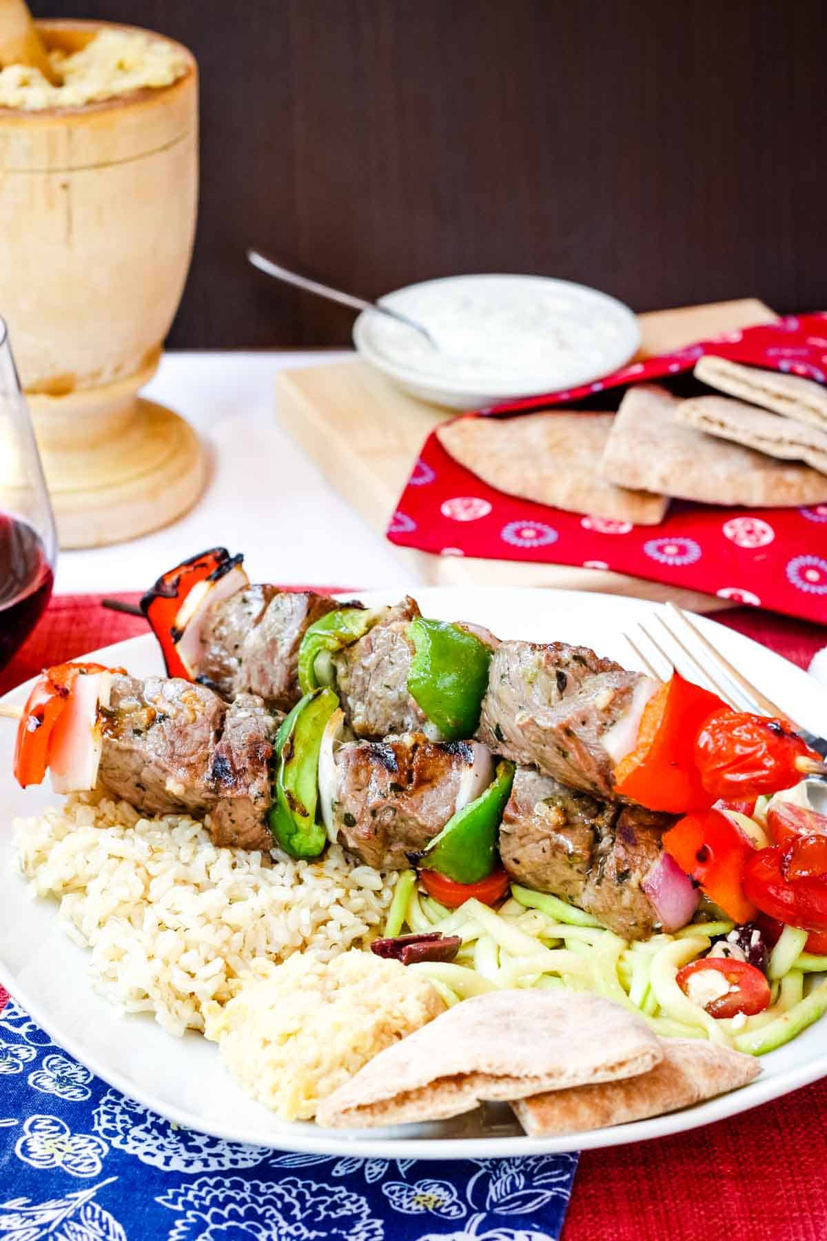 Grilled Greek Beef Kabobs on a plate with rice and pita with more pita on a cutting board with bowls of hummus and tzatziki.