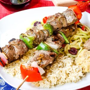 Two beef souvlaki kabobs on a bed of brown rice with hummus and a zucchini noodle salad.