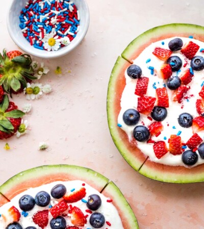 Overhead shot of two watermelon fruit pizzas topped with yogurt and berries plus a bowl of sprinkles