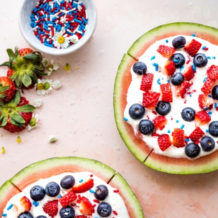 Overhead shot of two watermelon fruit pizzas topped with yogurt and berries plus a bowl of sprinkles