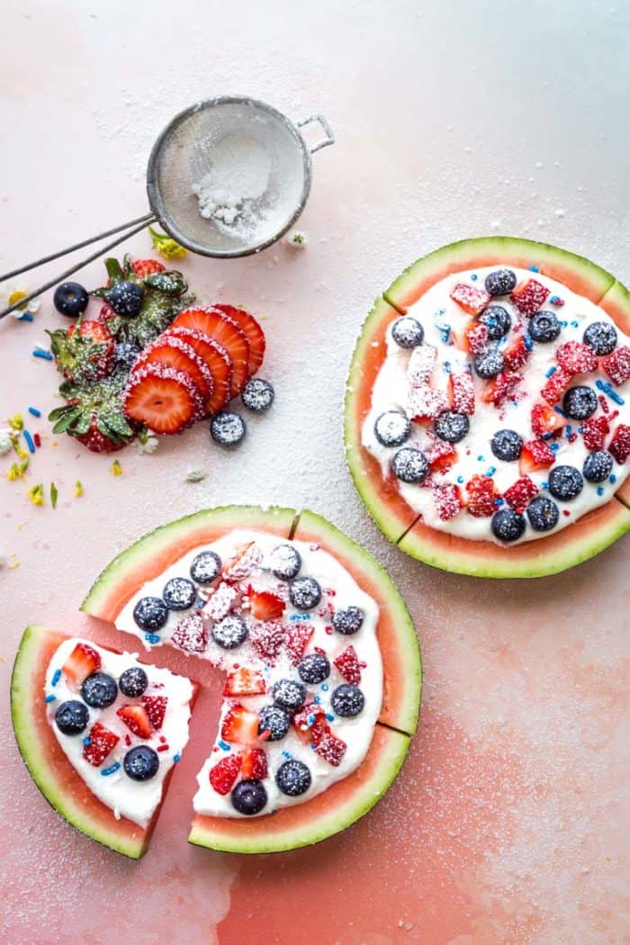 Two rounds of watermelon topped with whipped cream, berries, sprinkles, and powdered sugar and sliced like a fruit pizza