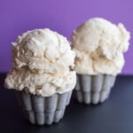 Two metals cups of cheesecake ice cream on a black tabletop with a purple background.