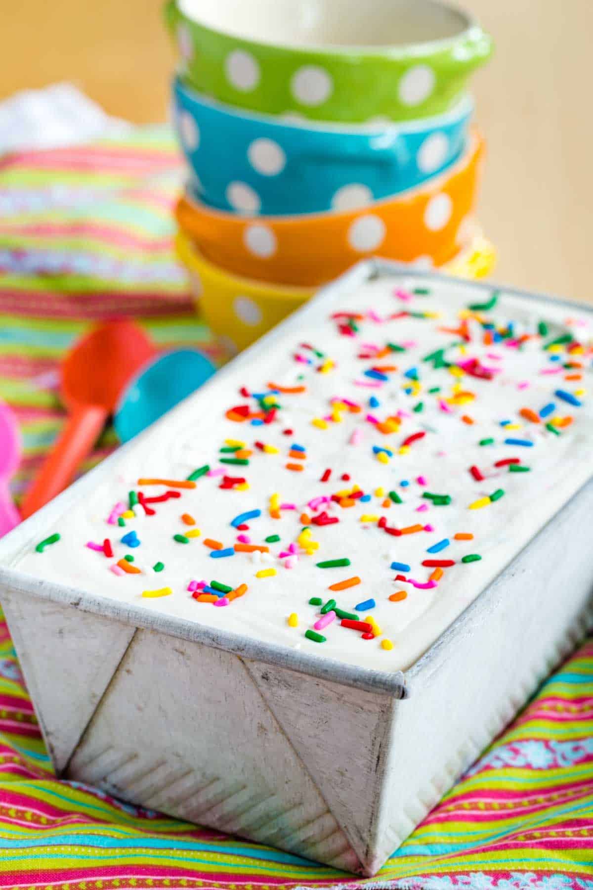 A batch of homemade Ice Cream with rainbow sprinkles scattered over the top in a pan with polka dot bowls in the background.