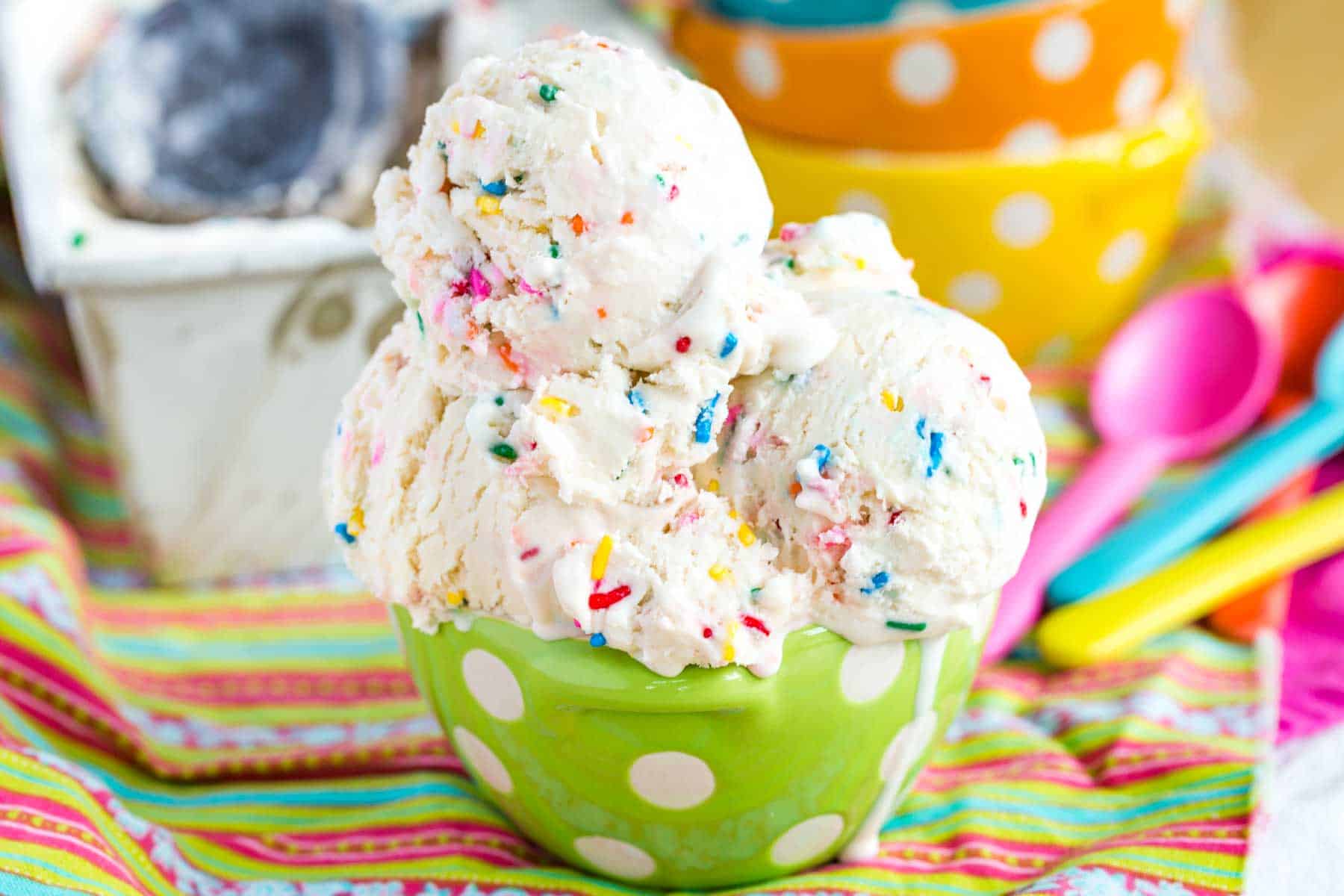Ice Cream filled with rainbow sprinkles in a green polka dot bowl with the ice cream container and more bowls and spoons in the background.