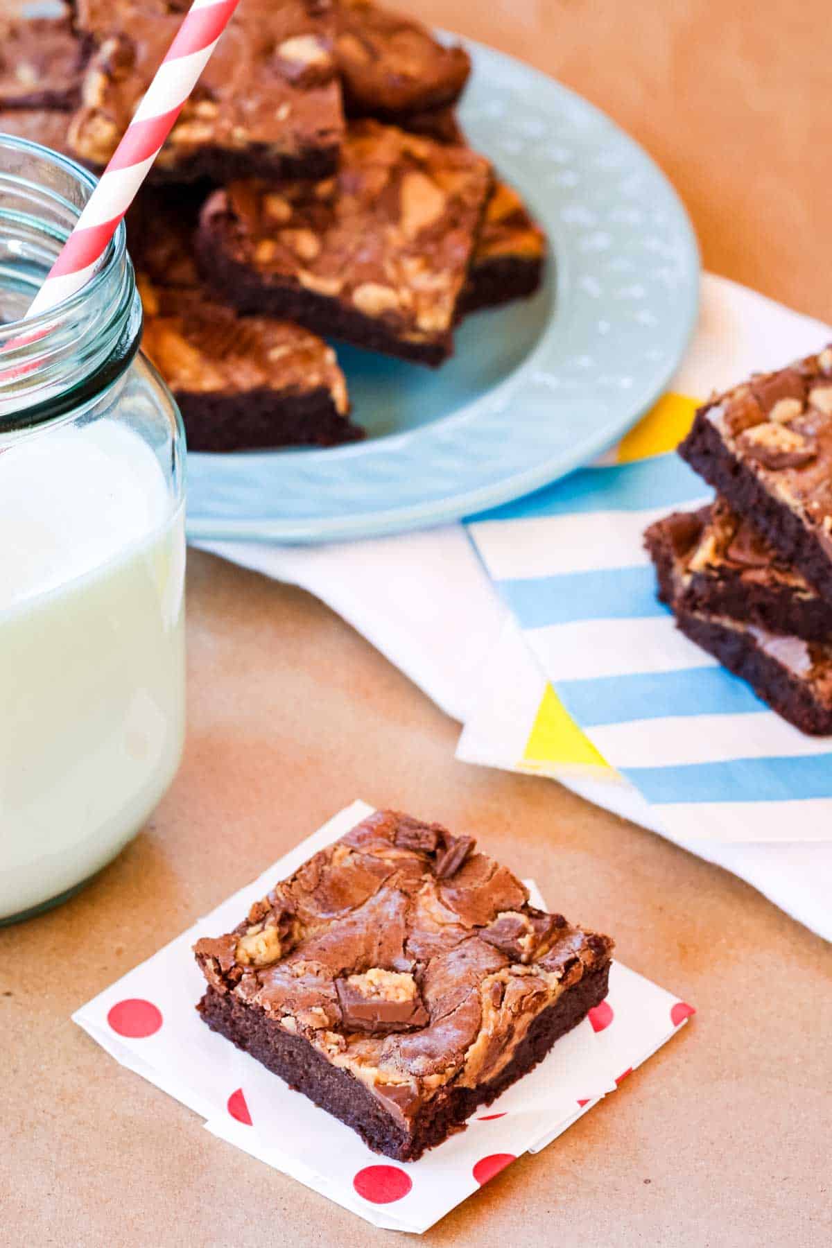 A peanut butter brownie on a white napkin with red polka dots, next to a glass of milk with more brownies on a plate in the background.