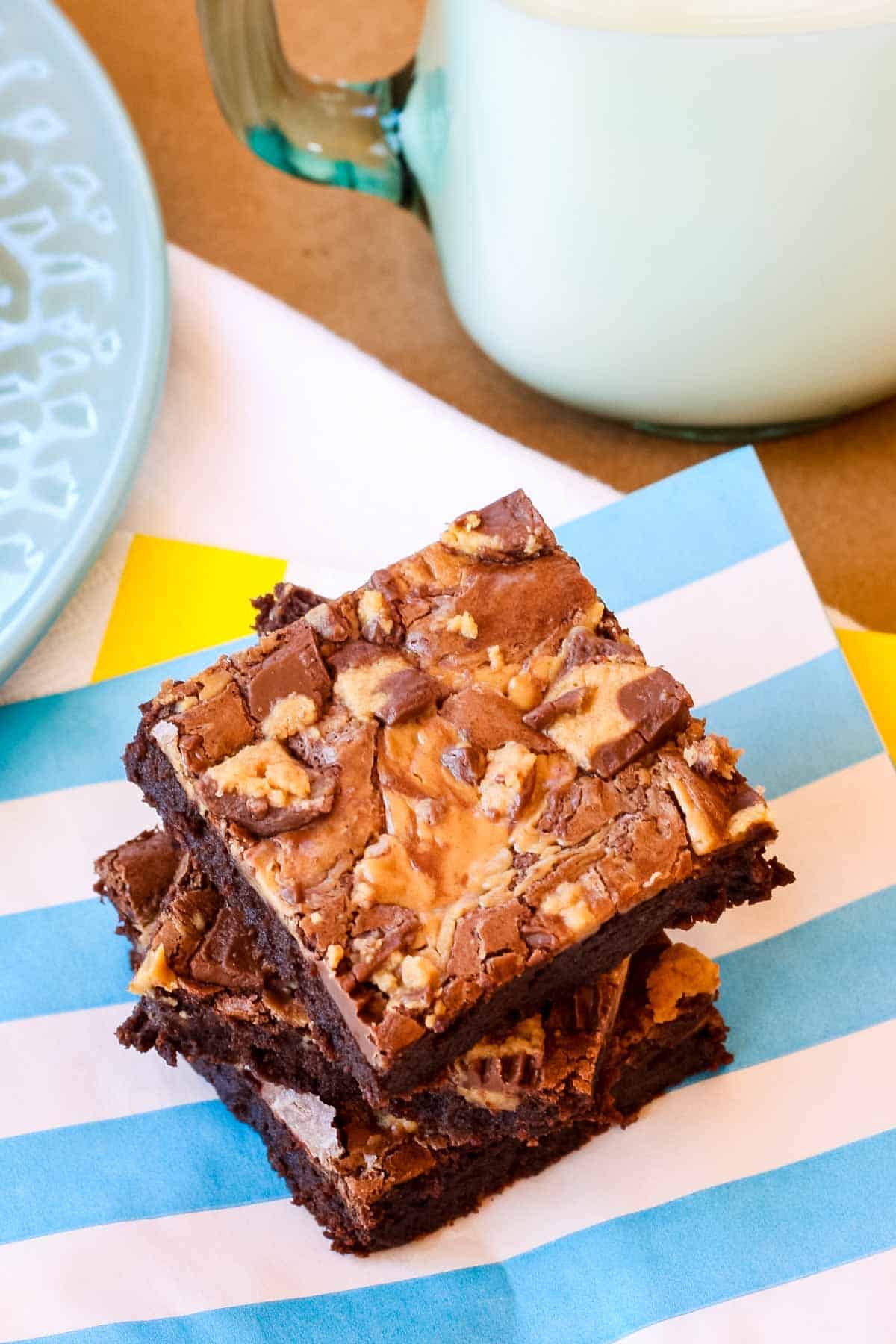 Three peanut butter brownies stacked on a blue and white striped napkin next to a glass of milk.