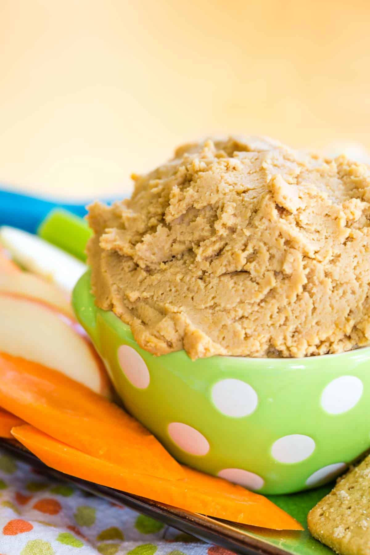 Peanut Butter Dessert Hummus in a green polka dot bowl with carrots and apples around it.