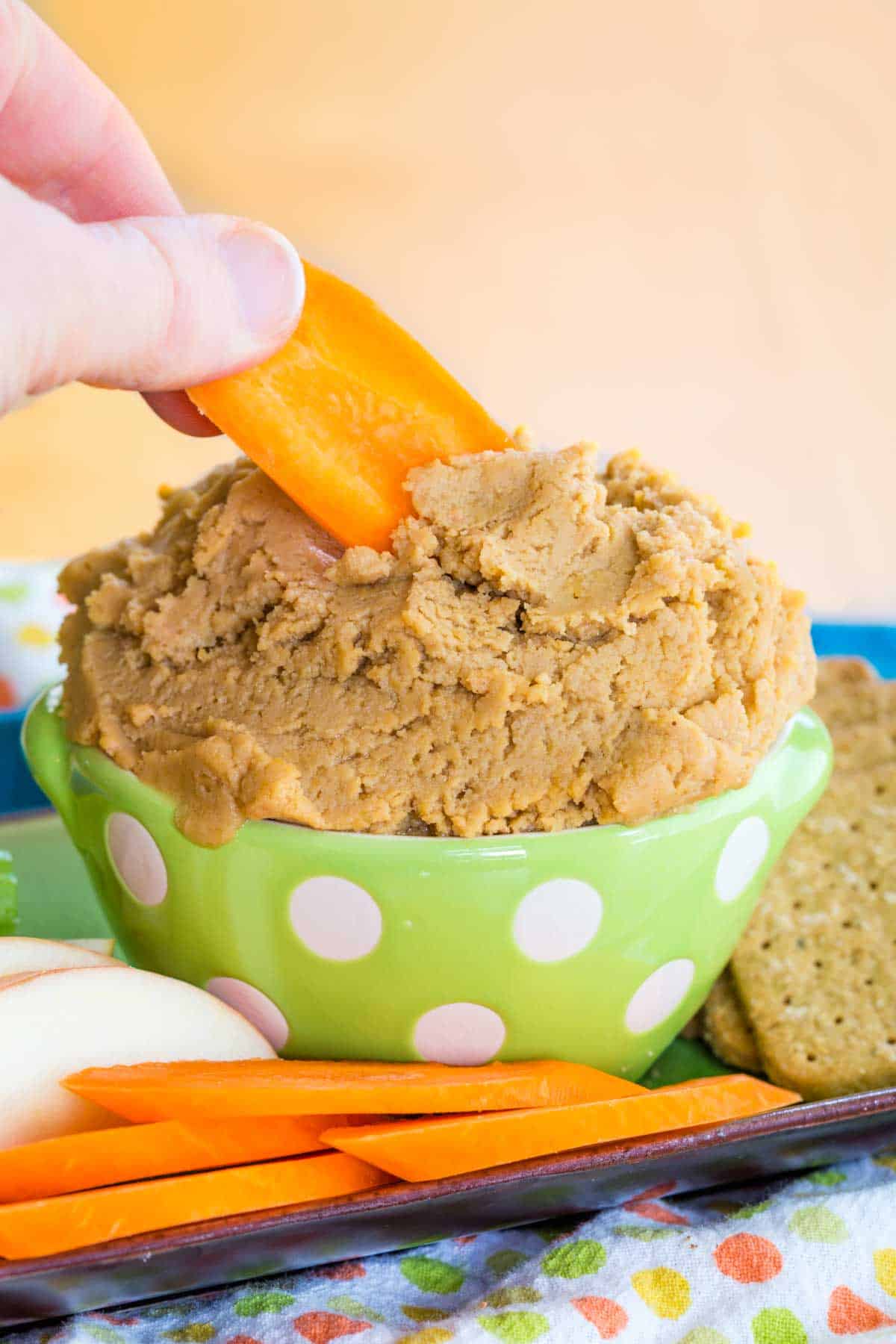 A hand dipping a carrot slice into a bowl of Sweet Peanut Butter Hummus.