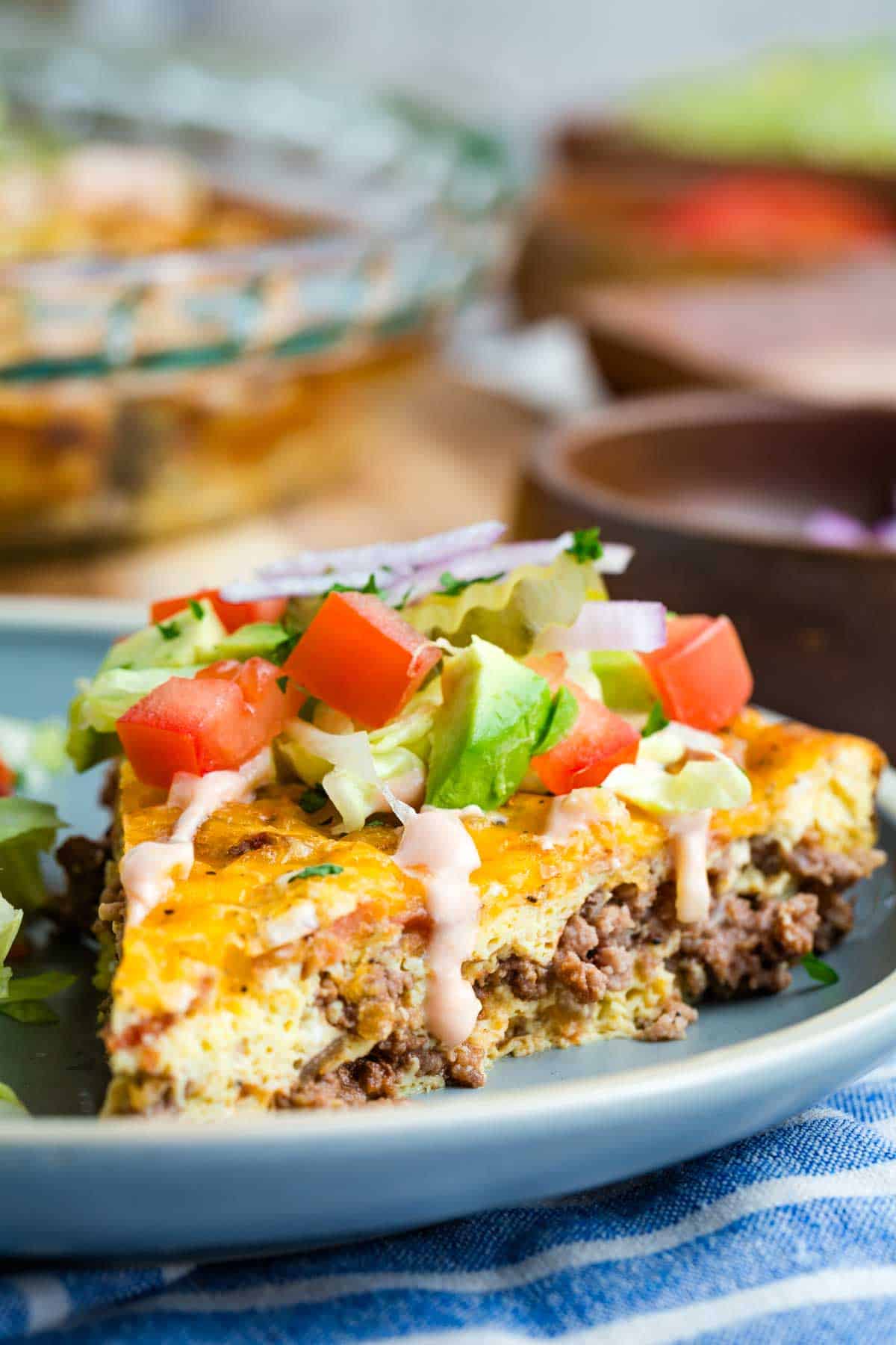 Bacon Cheeseburger Crustless Quiche is made with ground beef, bacon, and cheese, plus your favorite burger toppings. A slice of bacon cheeseburger crustless quiche on a blue plate