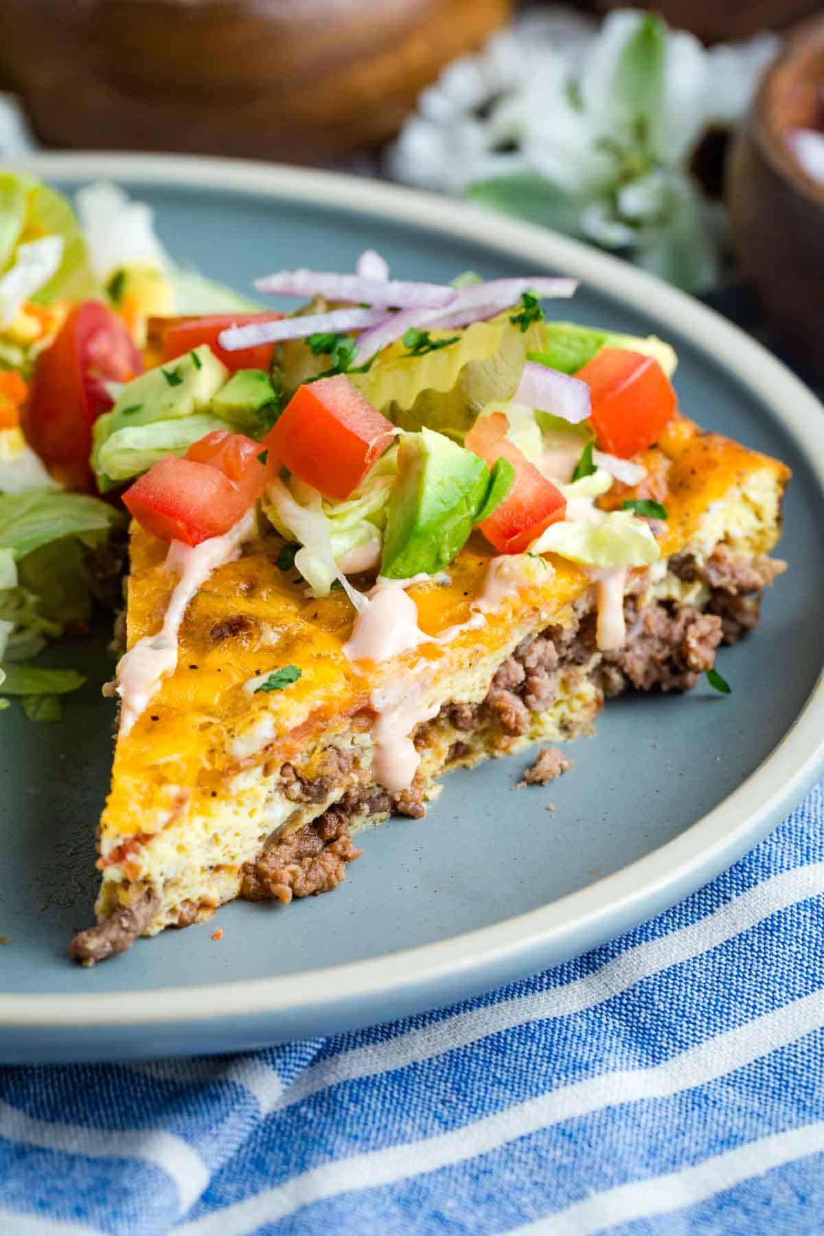Bacon Cheeseburger Crustless Quiche is made with ground beef, bacon, and cheese, plus your favorite burger toppings. A slice of bacon cheeseburger quiche on a blue plate