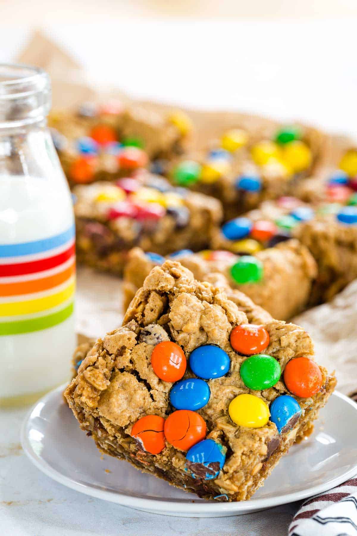 Monster Cookie Bars on a small white plate with one leaning on the other with more of the monster bars on a sheet of parchment paper.