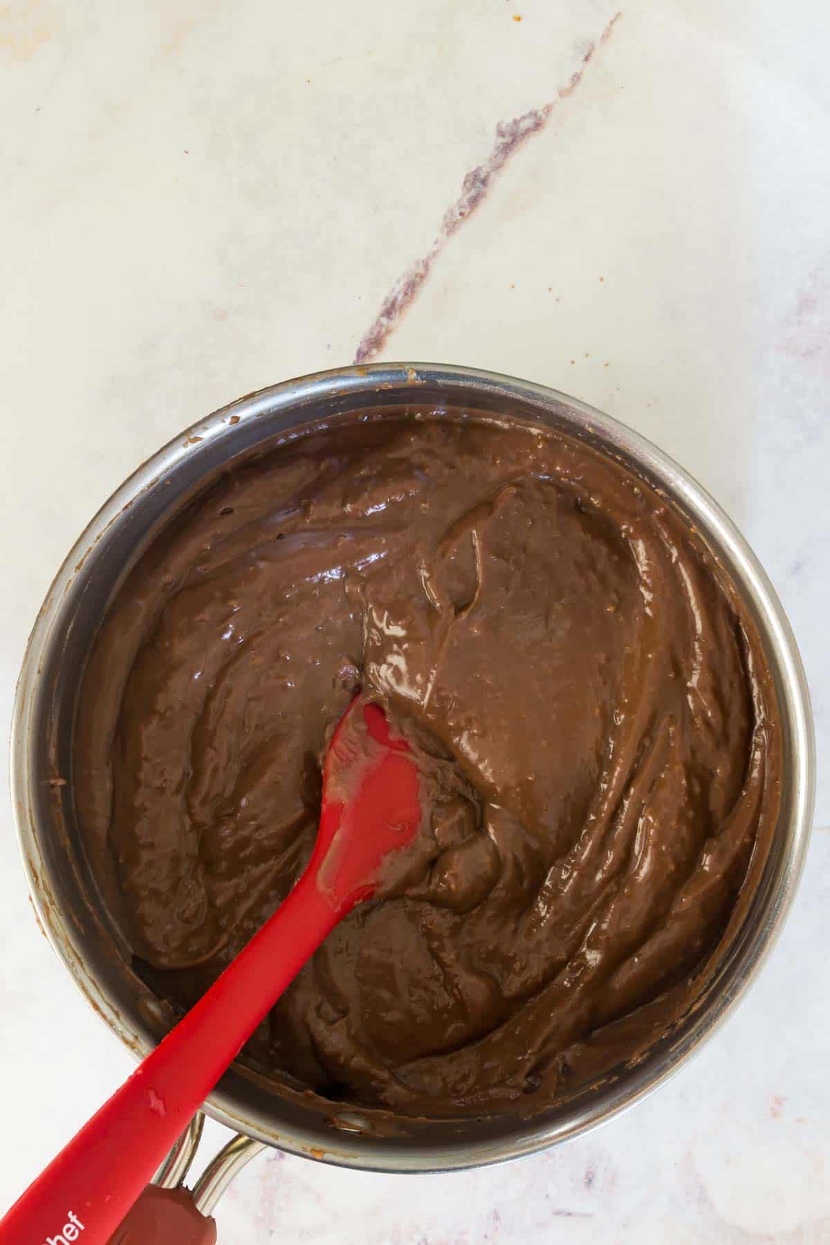 The chocolate pudding pie filling in a saucepan with a red stirring spoon.