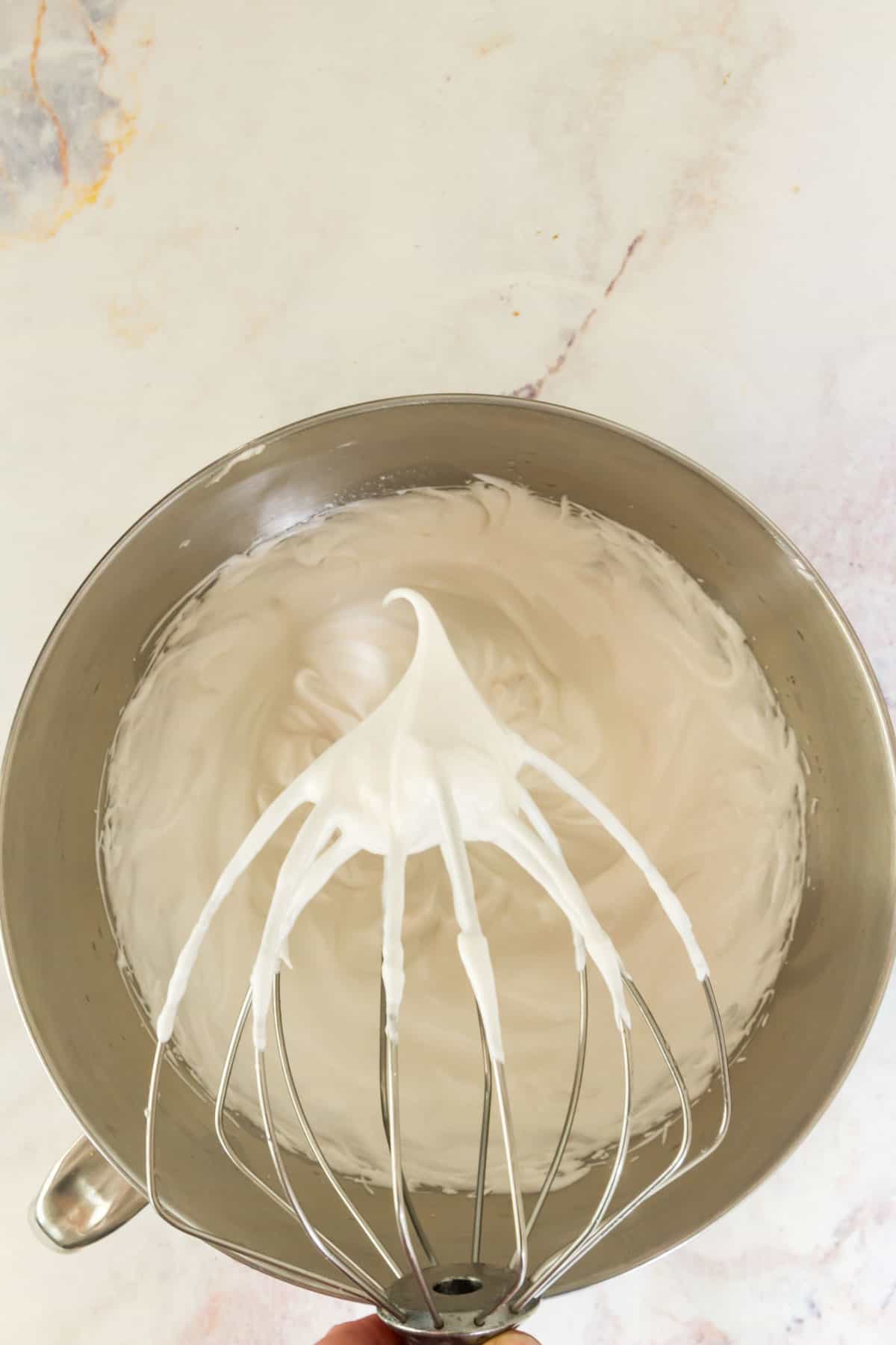 A stander mixer whisk attachment with a stiff meringue peak, with the bowl of the stand mixer in the background.