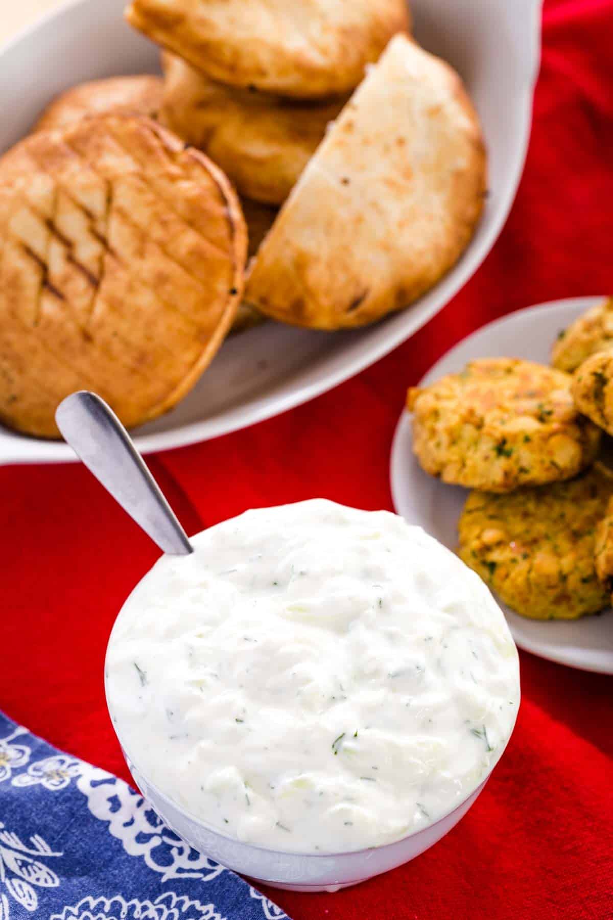 A bowl of Tzatziki sauce with pita and falafel on a red tablecloth.