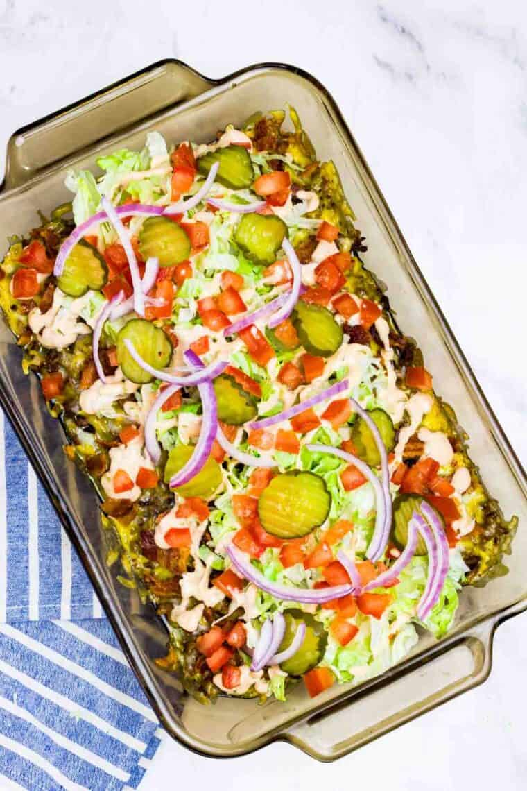A baking dish of bacon cheeseburger dip topped with lettuce, tomato, onion, and pickles