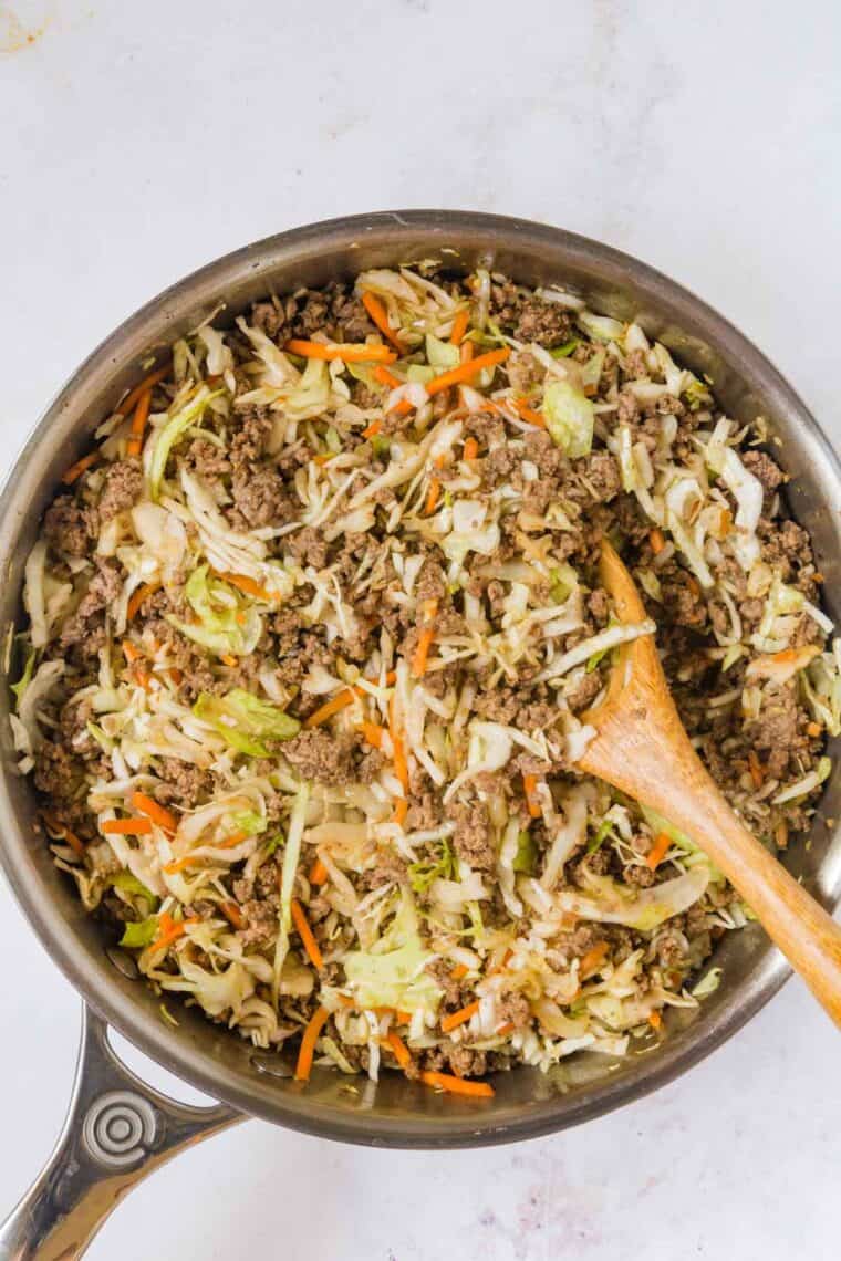 Ground beef and coleslaw mix combined in a pot, stirred with a wooden spoon.