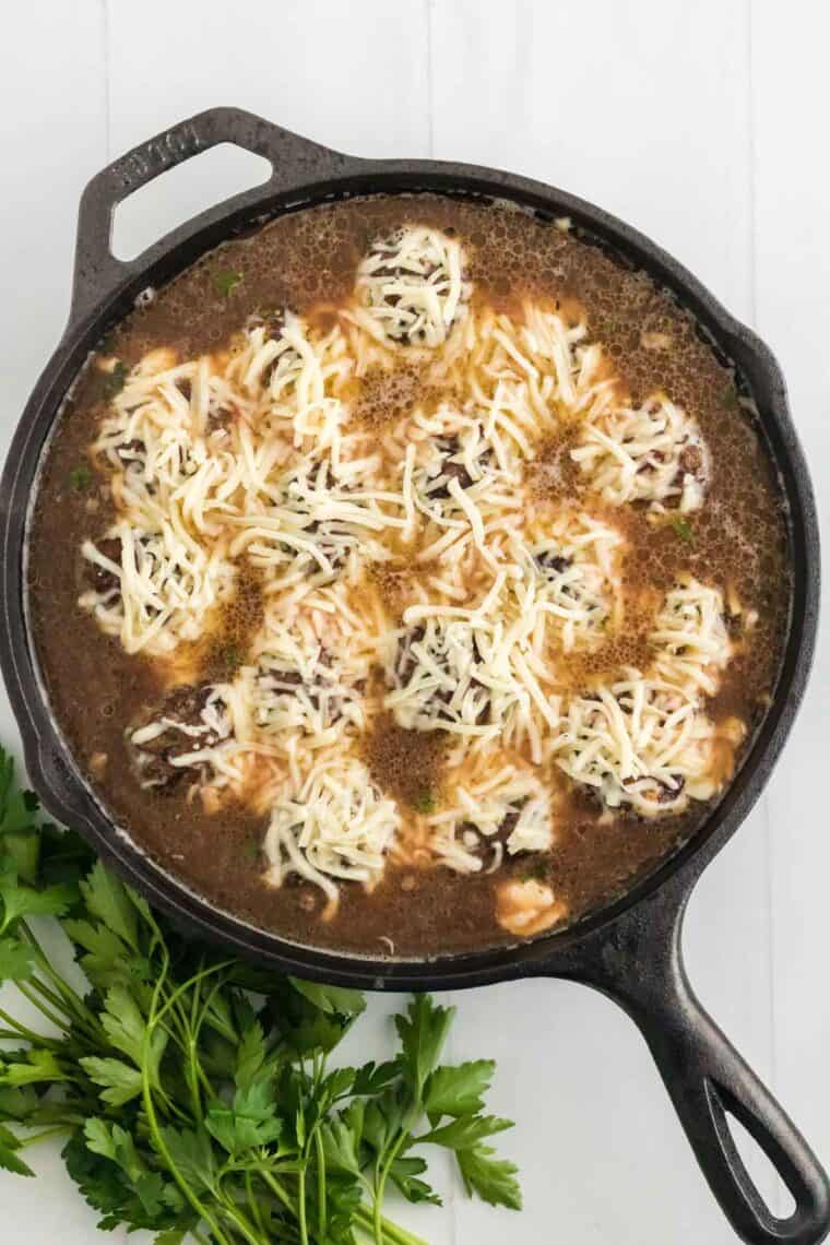Shredded cheese sprinkled over top French onion meatballs in a skillet.