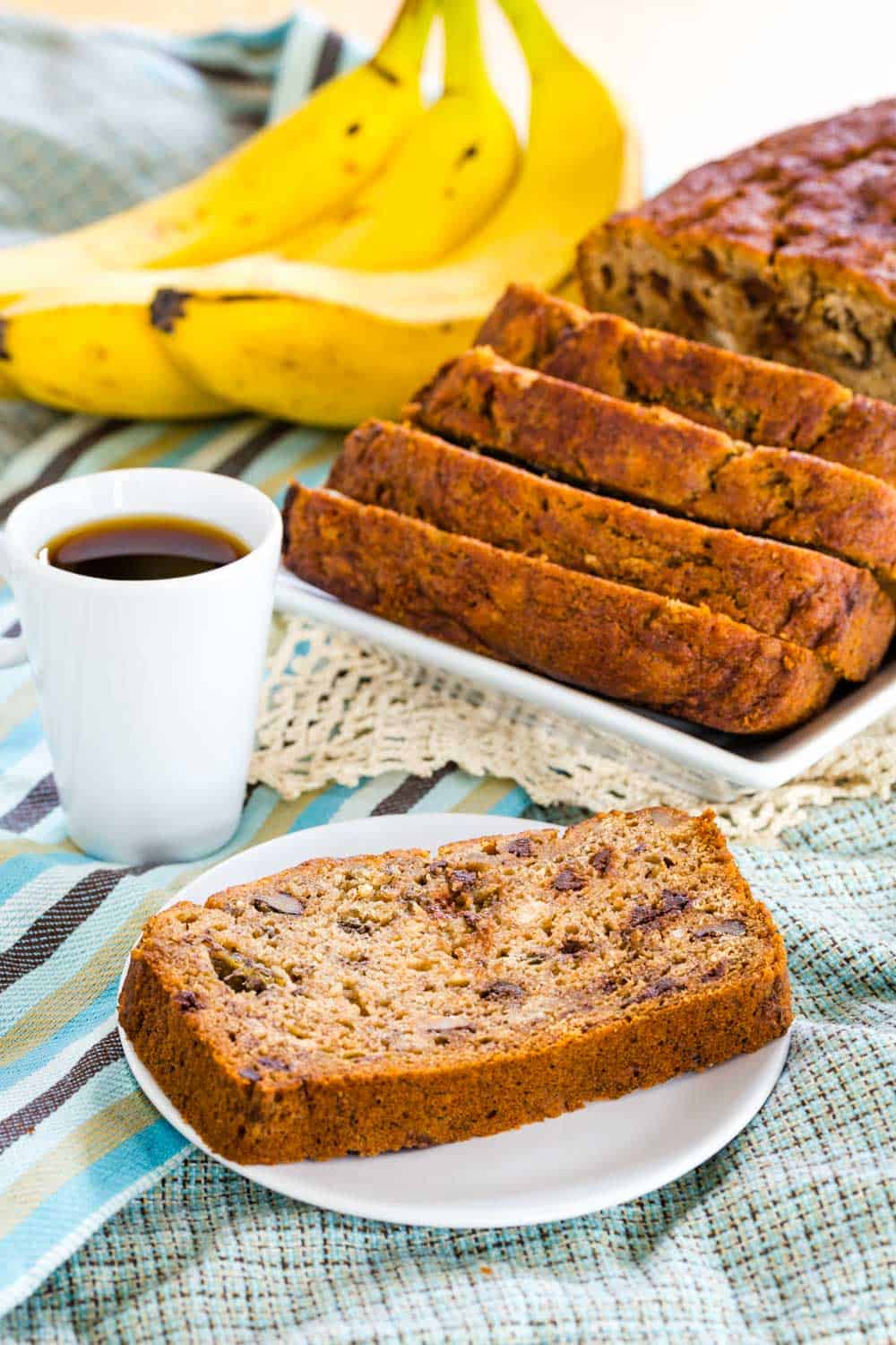 A piece of gluten free banana bread on a plate with a cup of coffee on top of striped cloth napkins.