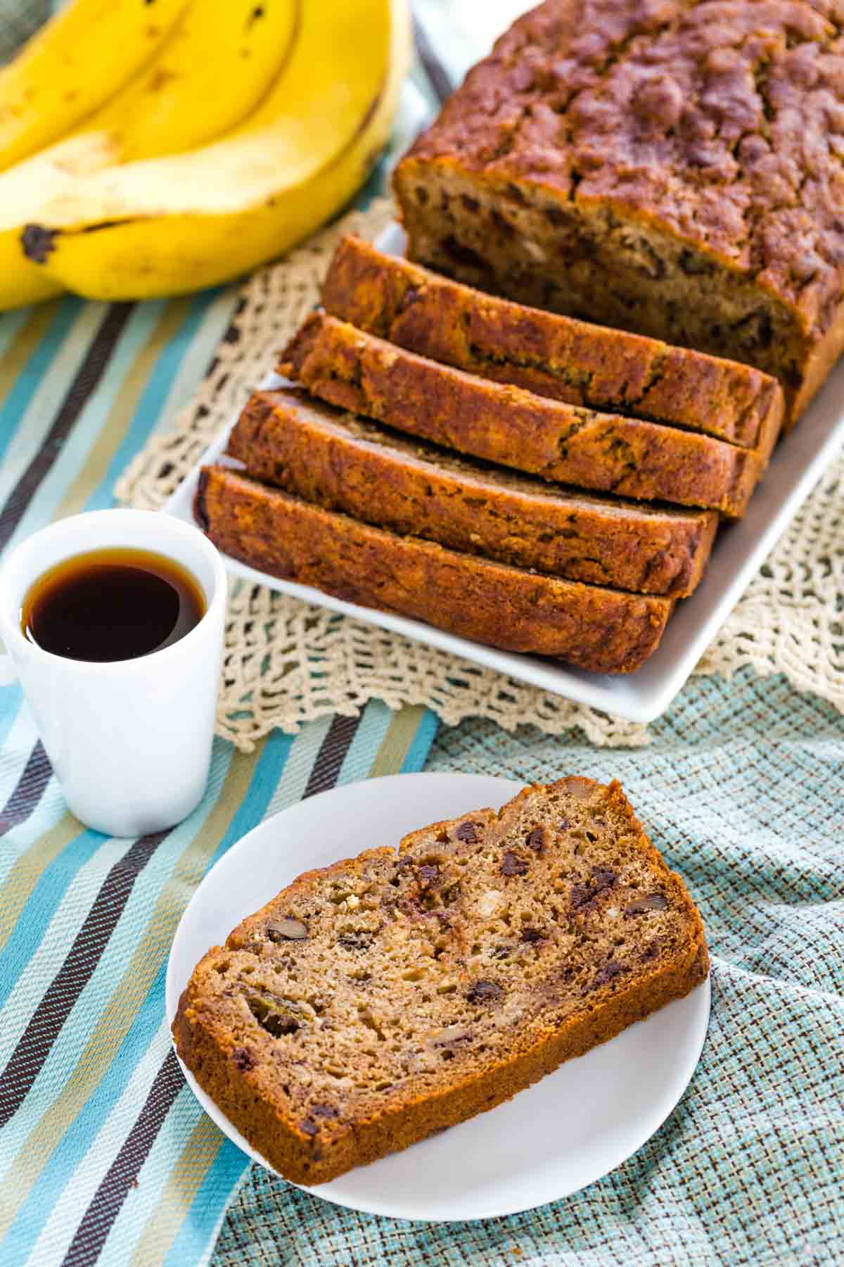 A sliced loaf of gluten free banana nut bread on a white rectangular plate on top of cloth napkins and placemats.