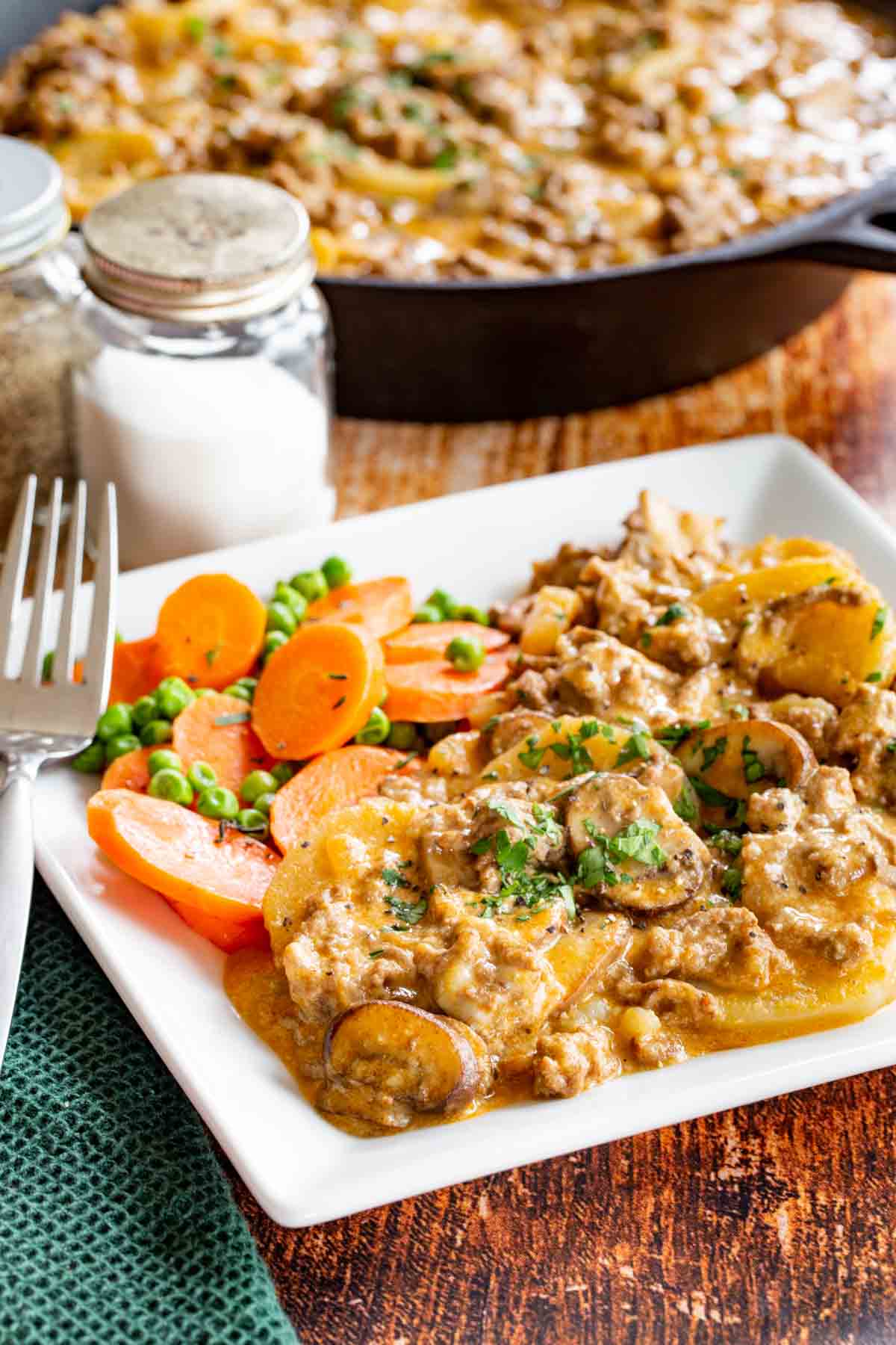 Hamburger Potato Skillet Meal Served on a white plate with peas and carrots in front of salt and pepper shakers and a skillet with the rest of the dish.