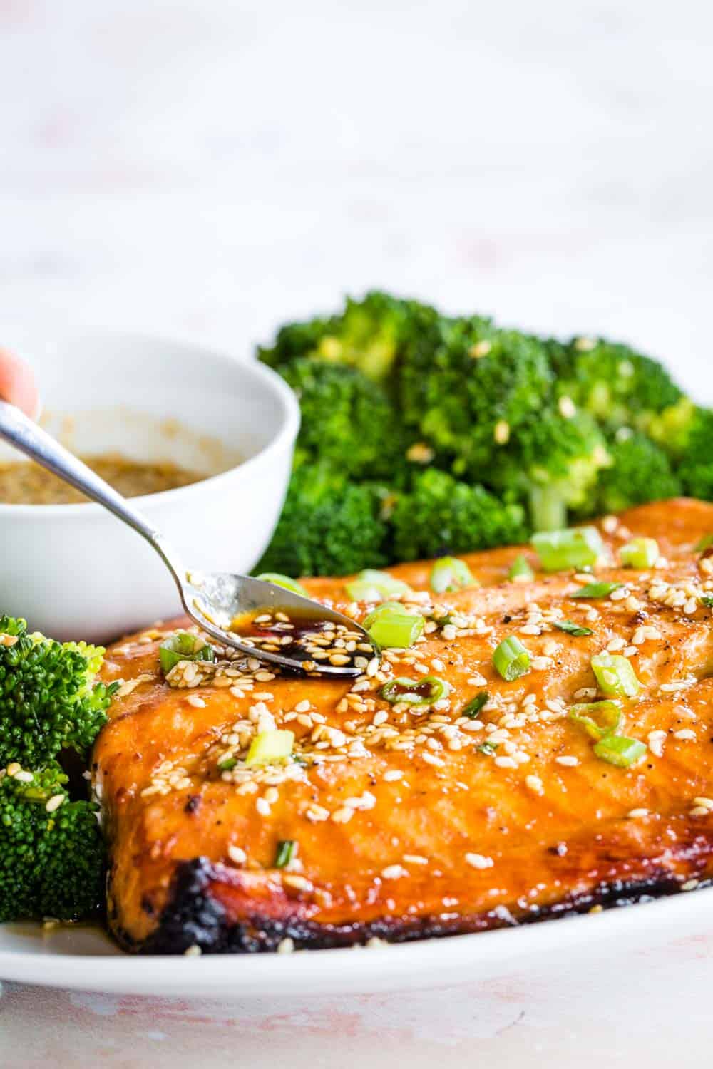 Spooning sesame maple glaze over broiled salmon on a plate with broccoli.