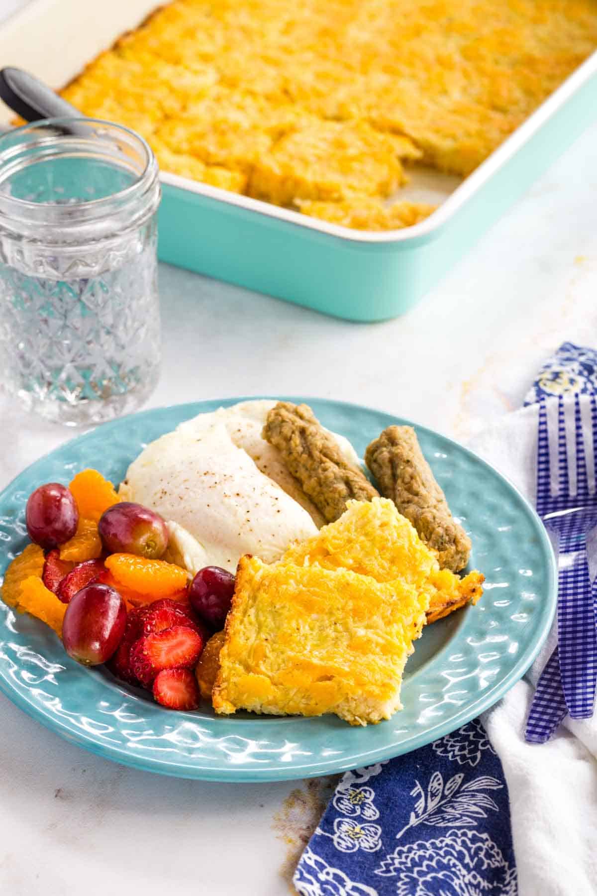 A plage of sausage and eggs with squares of cauliflower hash browns on a blue plate with the baking pan of the rest of the casserole behind it.