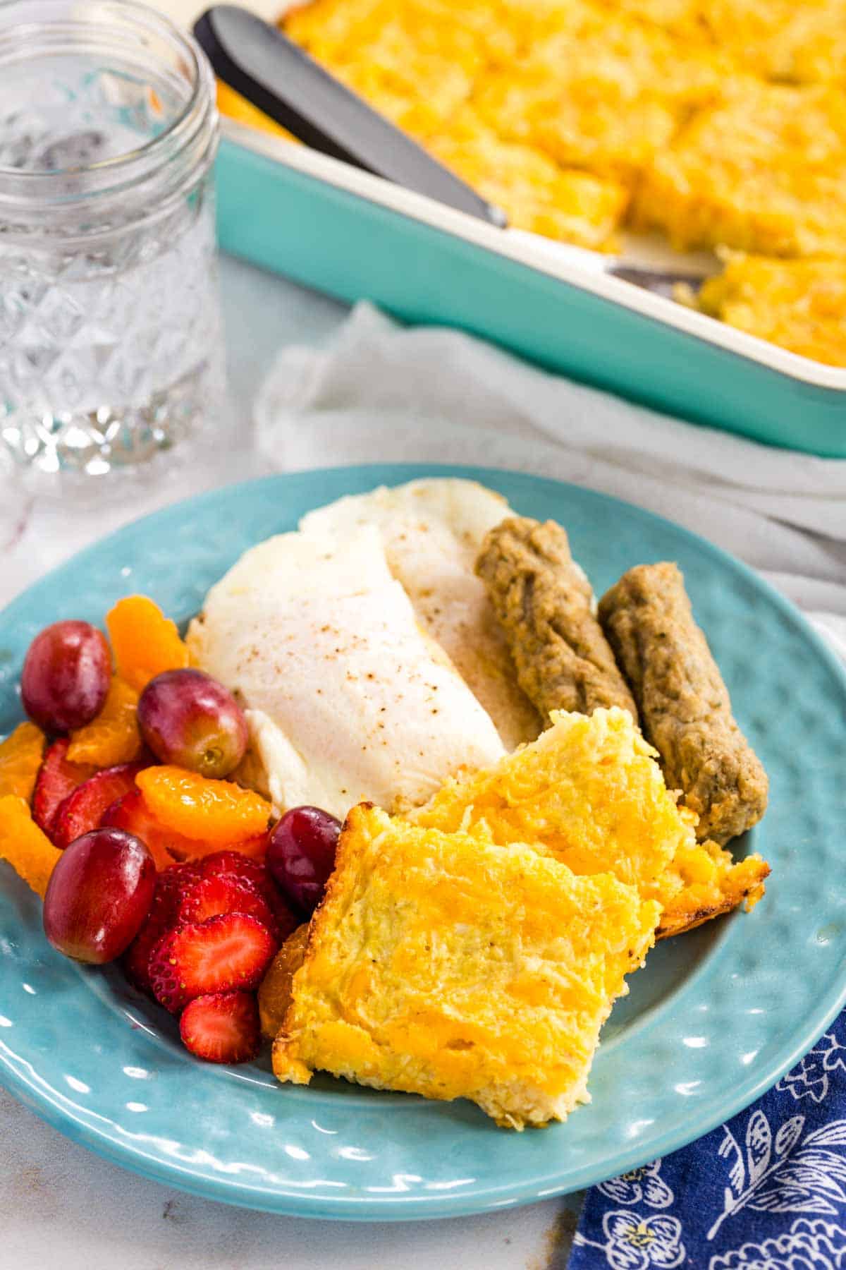 Squares of Cheesy Cauliflower Hash Browns Casserole on a plate with sausage, eggs, and fruit.