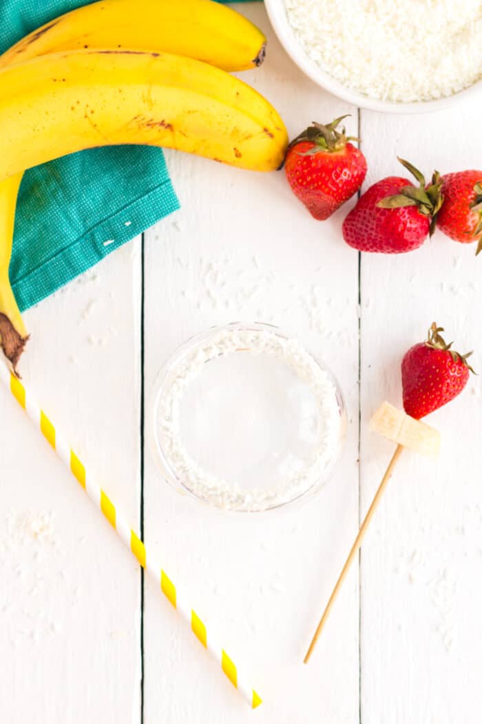 Coconut on the rim of a jar, a striped straw, and a strawberry and banana slice on a skewer to garnish a smoothie