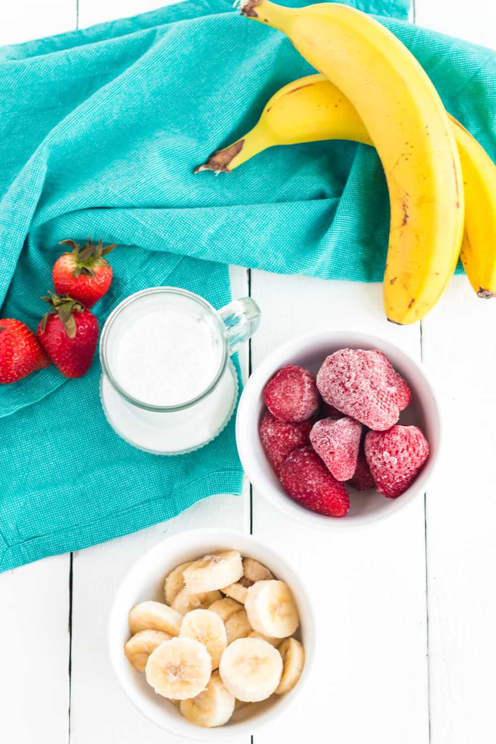 A pitcher of coconut milk, and bowls od frozen bananas, and frozen strawberries plus fresh fruit and a teal napkin on a tabletop.