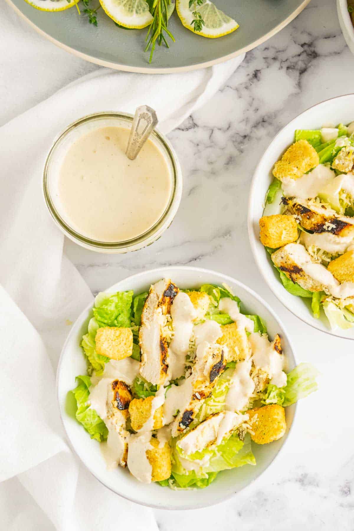 A jar of dressing and bowls of Chicken Caesar Salad