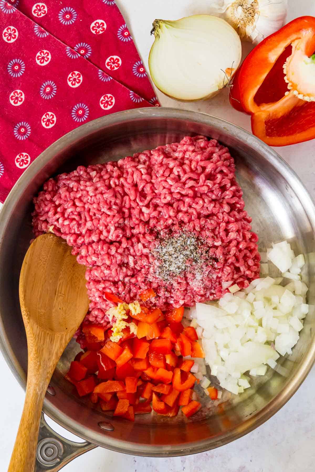 Raw ground beef with chopped onion and red bell pepper in a skillet with a wooden spoon.