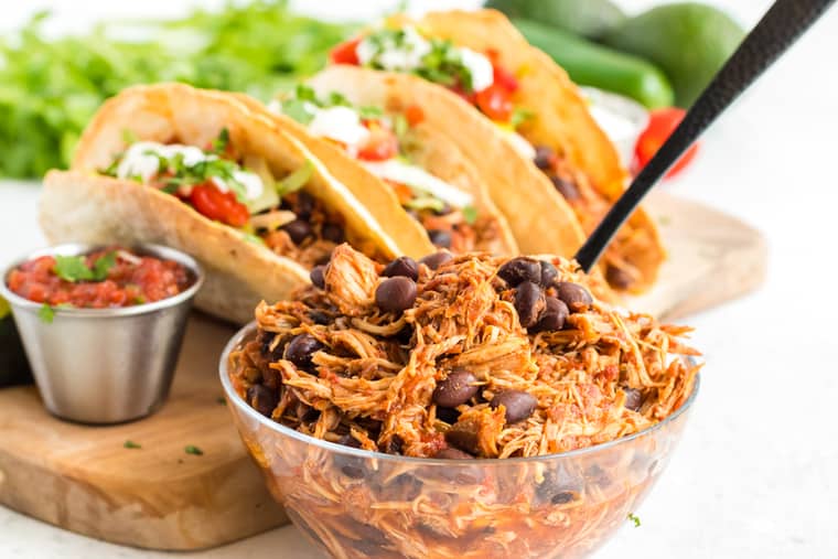 shredded chicken with black beans in a bowl with tacos in the background