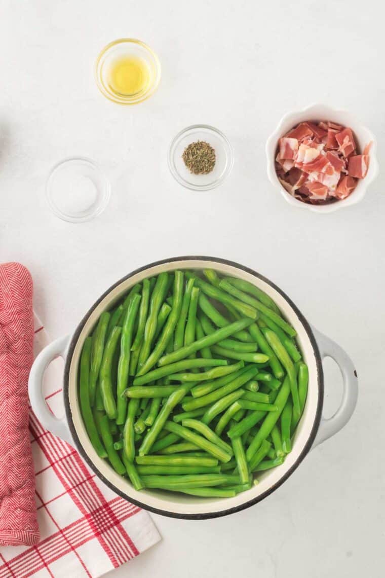 Green beans being boiled in a pot surrounded by bowls of the other recipe ingredients.